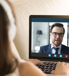 A woman wearing headphones is looking at a computer screen where she is having a video interview with a man who is wearing a suit.