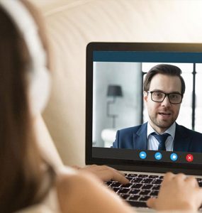 A woman wearing headphones is looking at a computer screen where she is having a video interview with a man who is wearing a suit.