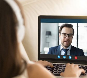 A woman wearing headphones is looking at a computer screen where she is having a video interview with a man who is wearing a suit.