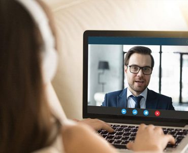 A woman wearing headphones is looking at a computer screen where she is having a video interview with a man who is wearing a suit.