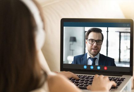 A woman wearing headphones is looking at a computer screen where she is having a video interview with a man who is wearing a suit.