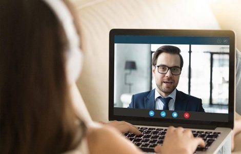 A woman wearing headphones is looking at a computer screen where she is having a video interview with a man who is wearing a suit.