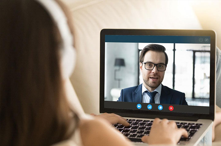 A woman wearing headphones is looking at a computer screen where she is having a video interview with a man who is wearing a suit.